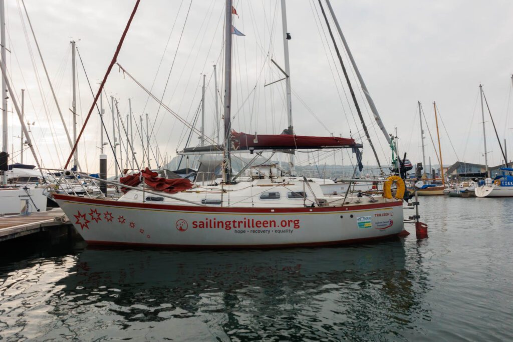 Image shows a grey boat in grey green water with a grey sky. the boat is left hand side towards the camera with bows to the left. There is red detailing on the sail boat including a logo reading sailingtrilleen.org