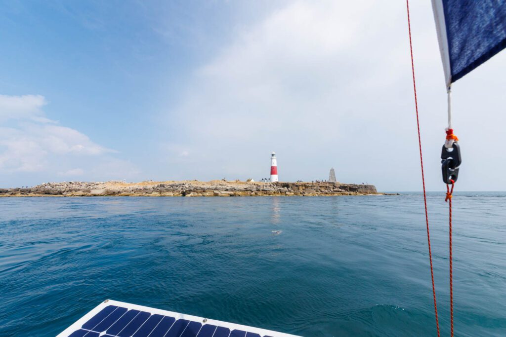 The image is bisected in half horizontally with a pale blue milky sky and cloud above and the a blue green calm sea below. along the divider line a low brown rocky headand extends from left to right. at the right hand end of the headland a red and white lighthouse stands tall. In the foreground are elements of boat rigging
