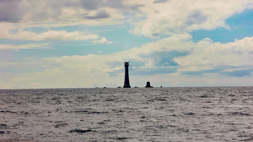 Divided into two parts, the lower sea, the upper a blue sky with complex cloud formations. On the horizon a silhouette of a lighthouse stands tall