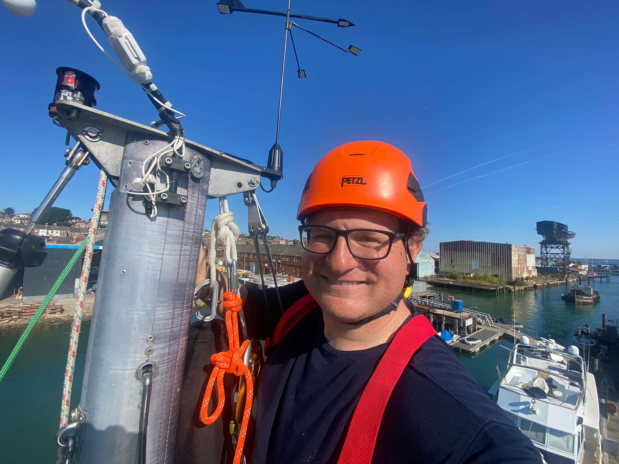 Ian is at the top of Trilleen's mast. He is wearing a orange helmet which stands out against a bright blue sky.