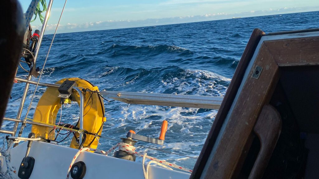 Shows a slight sea state with a canted horizon sloping downhill from right to left and blue sky above. In the foreground a lifebuoy is secured on Trilleen's guardrail 