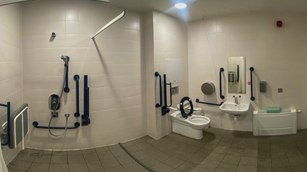 panorama of a good accessible toilet and shower facility. there is excellent contrast between the grey floor and bright which walls and the enough space around the toilet for a good transfer. The toilet is not a peninsula instal but the space would probably allow a right hand as well as left hand transfer.
