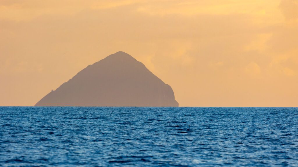 The conical shape of Ailsa Craig rises out of a smooth sea with an auburn skiy
