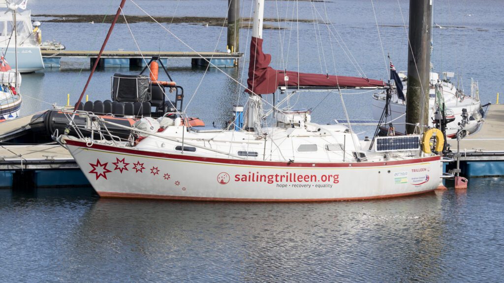 Shows a grey and red sailboat alongside a pontoon held in place by large steel piles. The sailboat has seven stars on the bow and text reading 'sailingtrilleen.org' on the side
