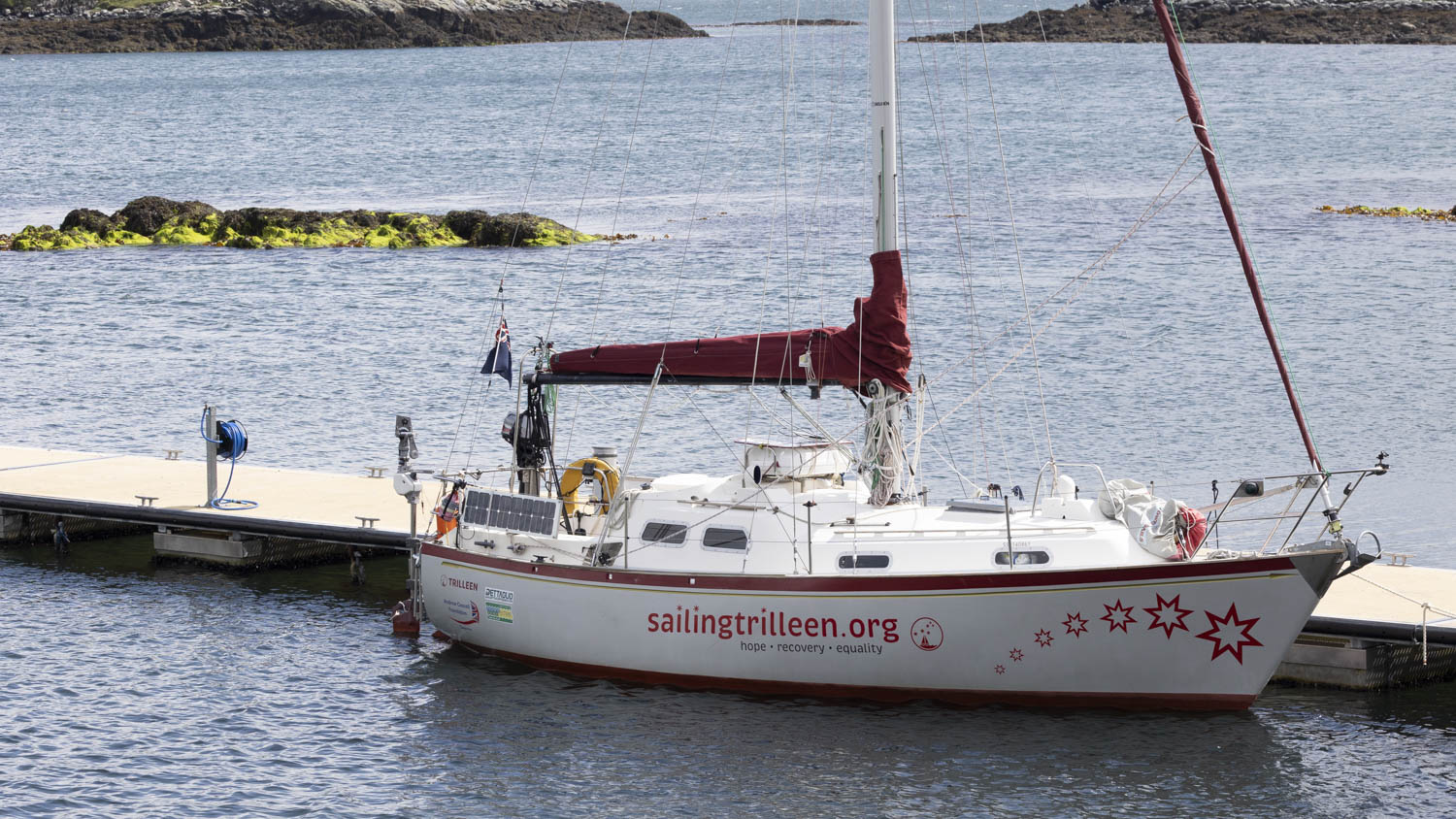 A sailboat marked SailingTrilleen with a red boom cover sits alongside a pontoon