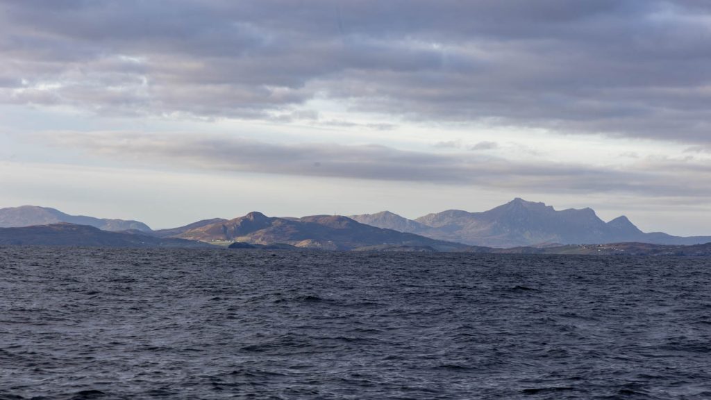 Blue fringed steep mountains lie on the horizon of a wind blown sea with a blue and clear sky above. The whole image is a gradient of blues