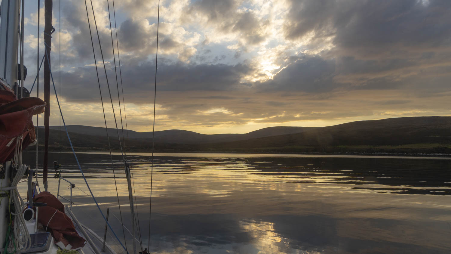 A boat's rigging and the front of her hull is visible in the foreground of a mirror smooth loch.c The loch is fringed by hills in deep shadow and there is a bronze and gold sunset visible through scattered clouds which is reflected in the water