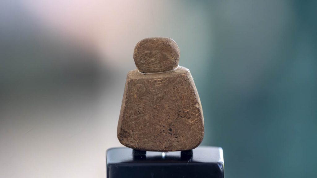 A sandstone figurine about 41mm high with an elliptical head set on a trapezoidal body is sat on a plinth against a blurred pink and green background.