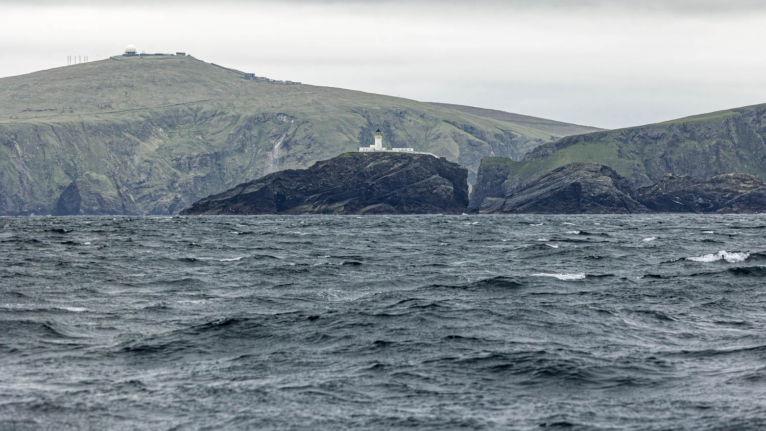A lighthouse stands out over a stormy sea with a white radome of a northern flank radars station on the cliff above.