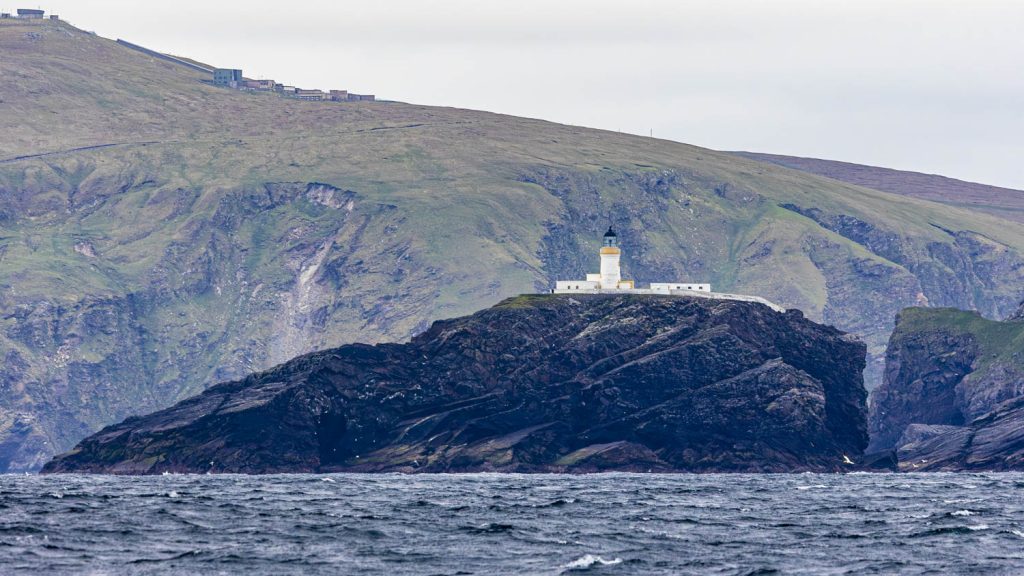 Muckle Flugga lighthouse rises on a sheer dark rock out of the ocean with the high cliffs of Unst and Saxa Vord rising behind.