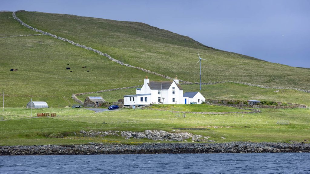 A White House stands with a wind turbine on the green sward of Unst. A stone fence leads diagonally up the hill