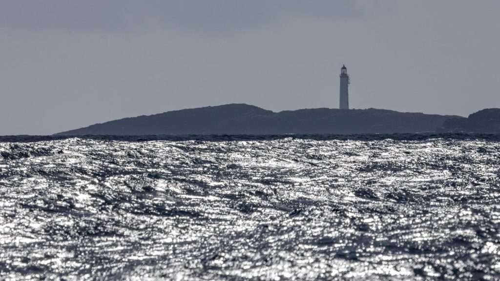 A lighthouse, backlit in the sun, stands over a low rocky blue islet and across a silvered sea.