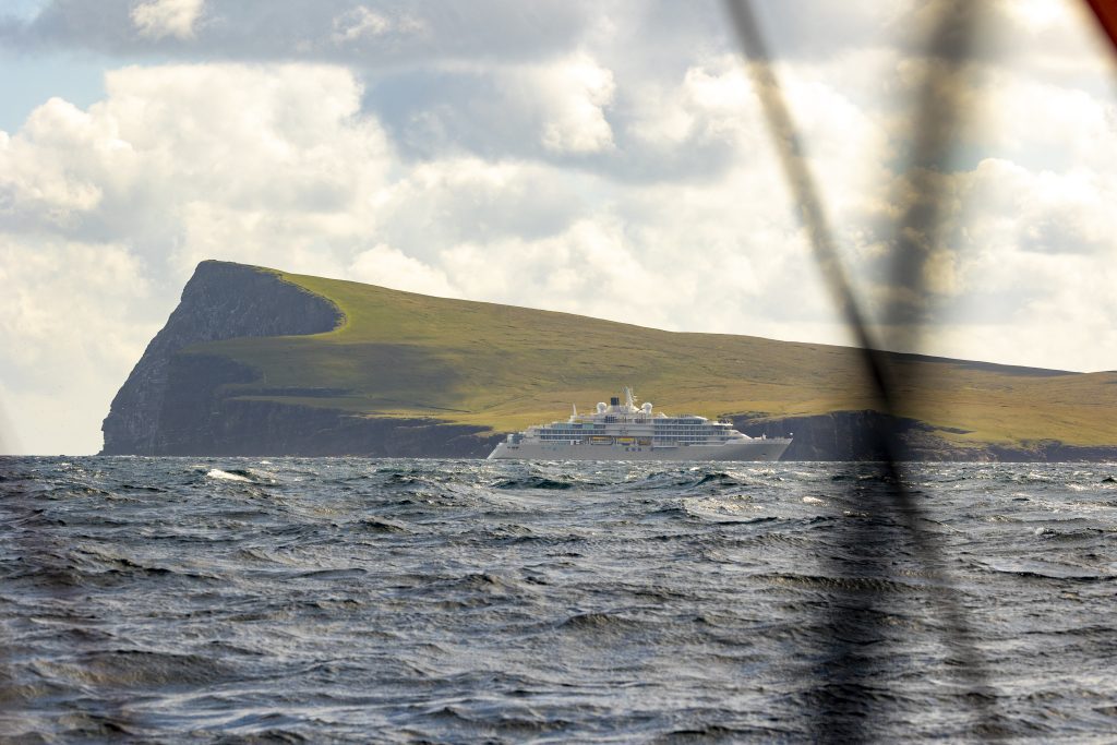 The great triangular profile of Noss Head towers above a small white and silver cruise ship: Silver Endeavor. The sea is a grey, blue with clearly visible white crests
