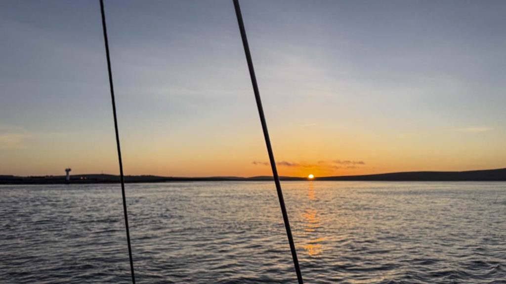 The sun, a flaming ball rises above the low horizon of Scapa Flow, a vast sea contained by the Orkney islands. The sky is graded from a milky blue to a violent orange and this is reflected in the water.