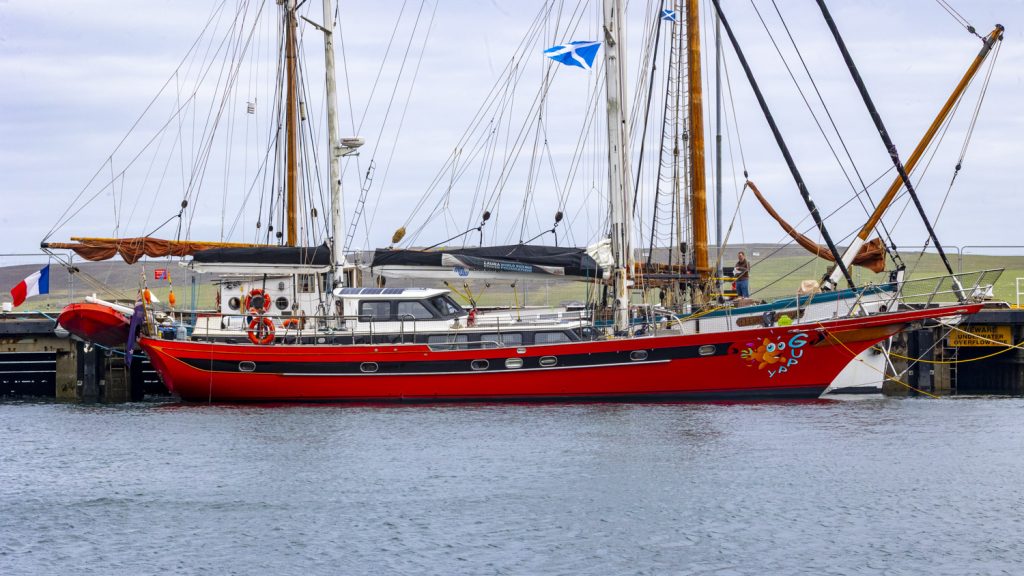 A bright red hulled seventy foot ketch lies outboard of an even bigger white hulled yacht in Lerwick