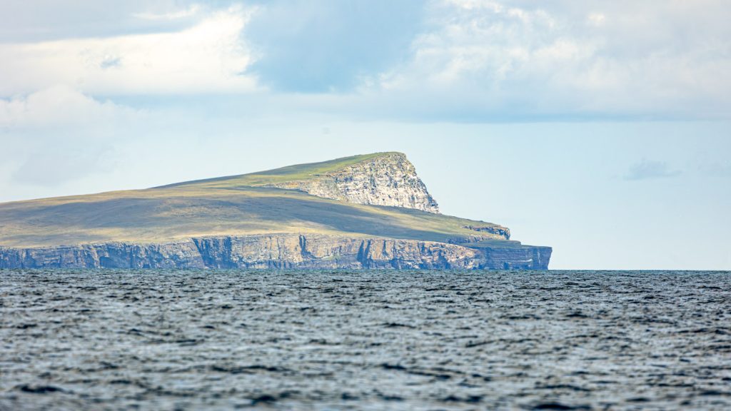 A vast triangular bulk of a headland sweeps steeply down to the sea. The shallow face of the headland is coated with fine green grass and the sky is a pale blue with fine high cloud