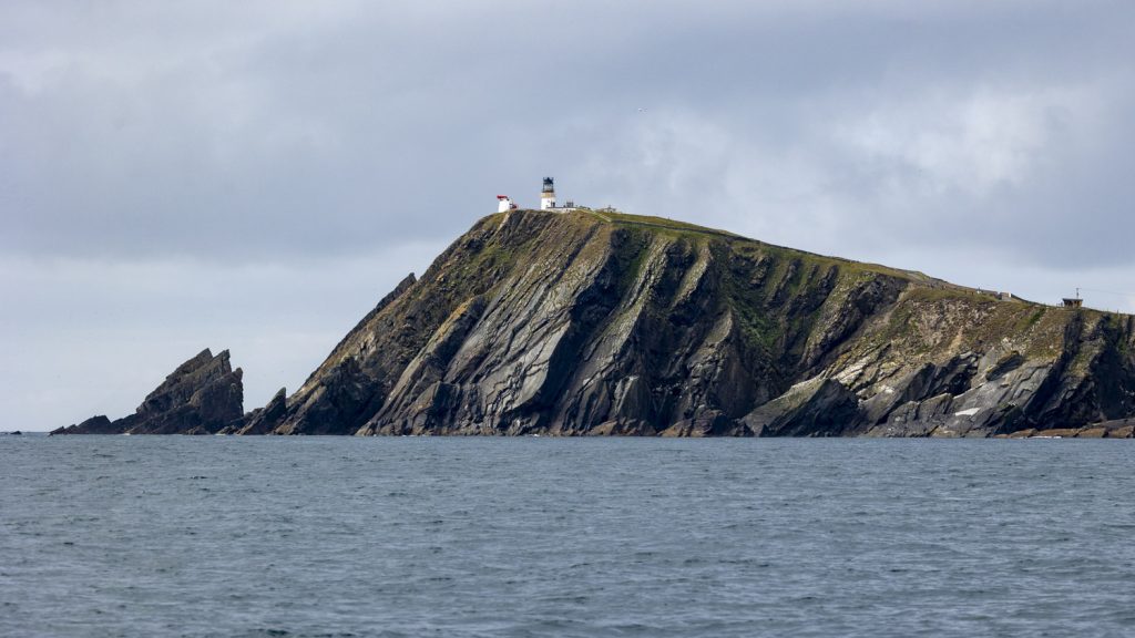 Steeply bedded planes of rock dive into a pale blue se. The sky behind the cliffs are low hanging altostratus. and a lighthouse pierces on the narrow grass strip at the clifftop