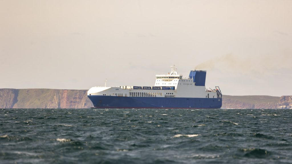 A blue and white cargo ship loaded with roll on roll off trailers cruises across a white capped sea past the imposing cliffs of the north shore of Scotland