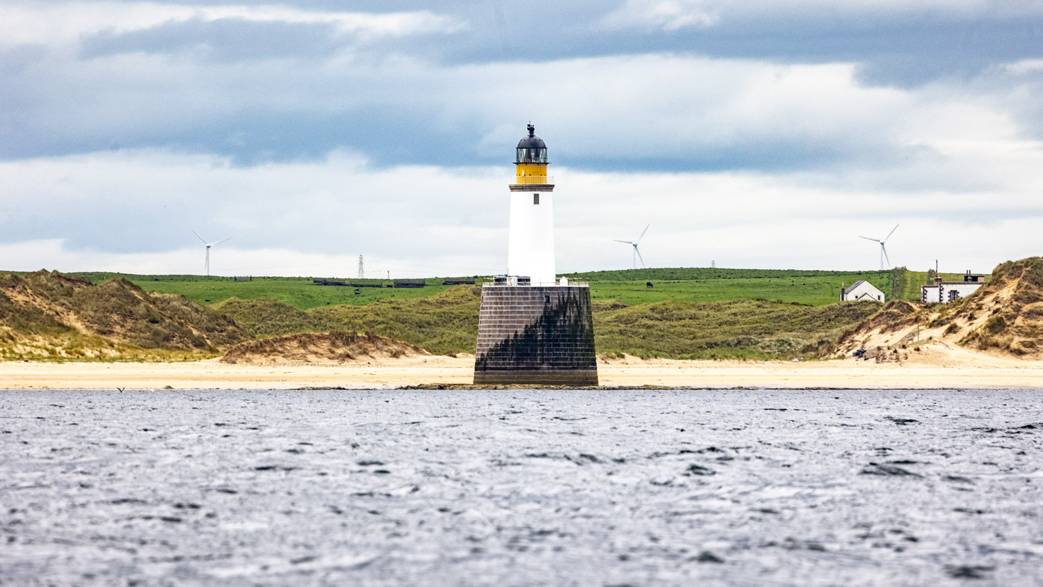 A low lighthouse with a yellow top, on a white pillar rises out of the sea in front of yellow dunes topped with marram grass