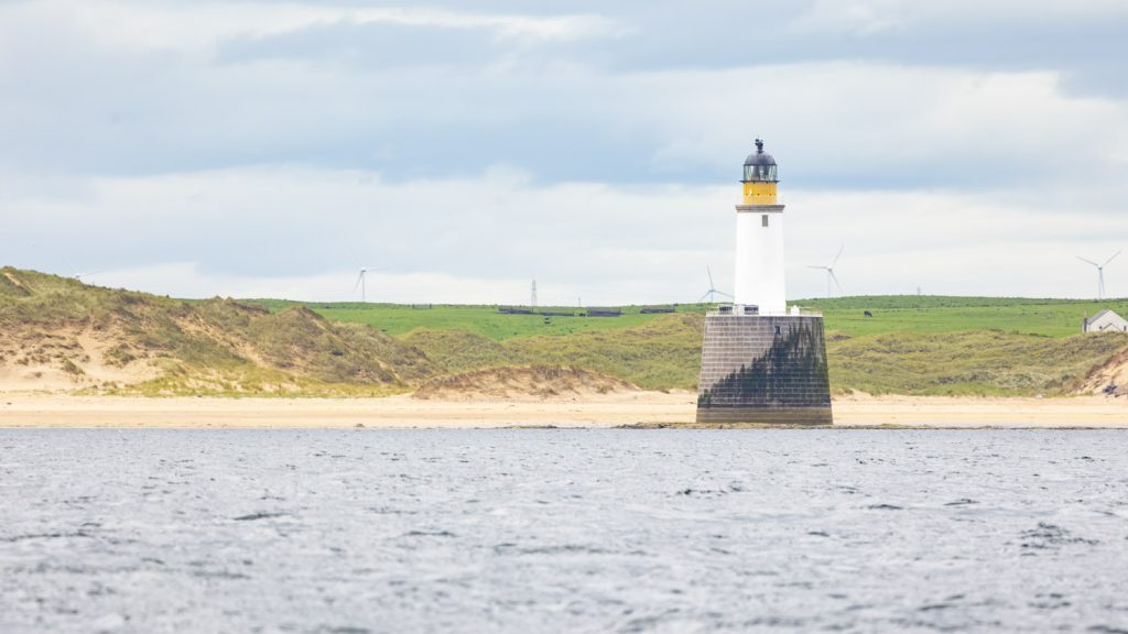 A low lighthouse rises out tof the sea in front off bright yellow sand dunes