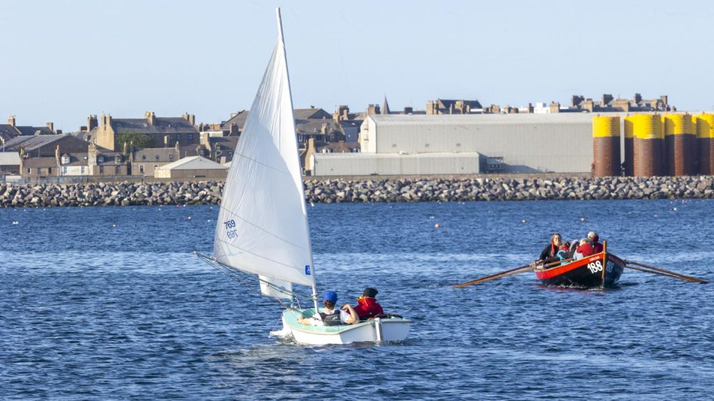 A disability adapted sailboat (Hansa) powers away downwind out of a marina towards an industrial port scene